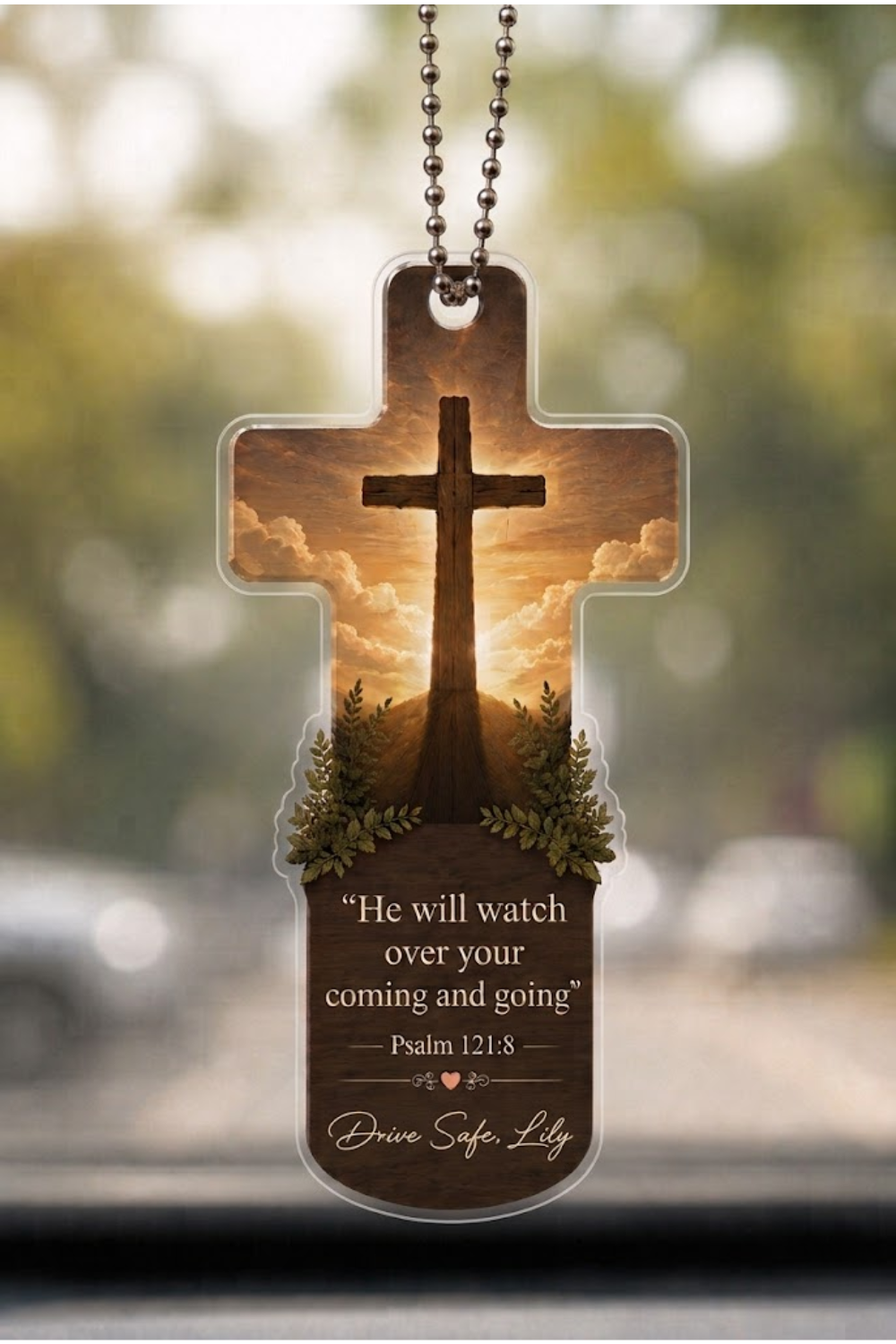 A cross-shaped car window charm with a printed sunset scene, a large cross silhouette, small floral details, and the text 'He will watch over your coming and going' Psalm 121:8 and 'Drive Safe Lily' at the bottom, hanging from a metal chain.