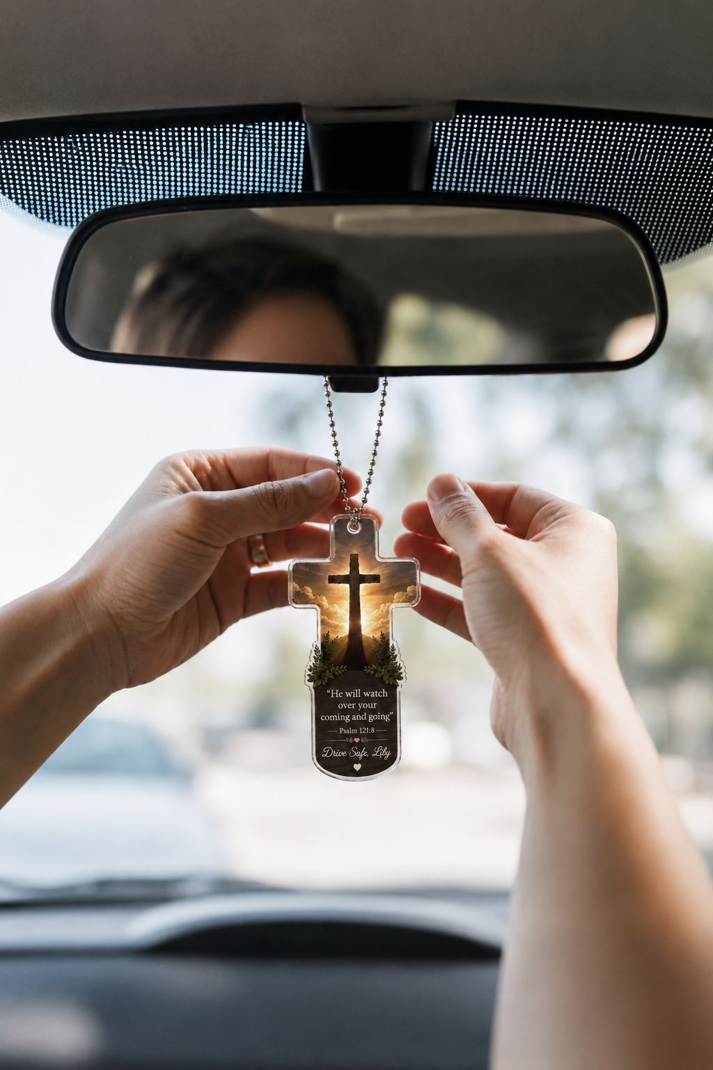 A person holds a clear cross-shaped car air freshener with a printed cross and Bible verse, positioned in front of a car rearview mirror.