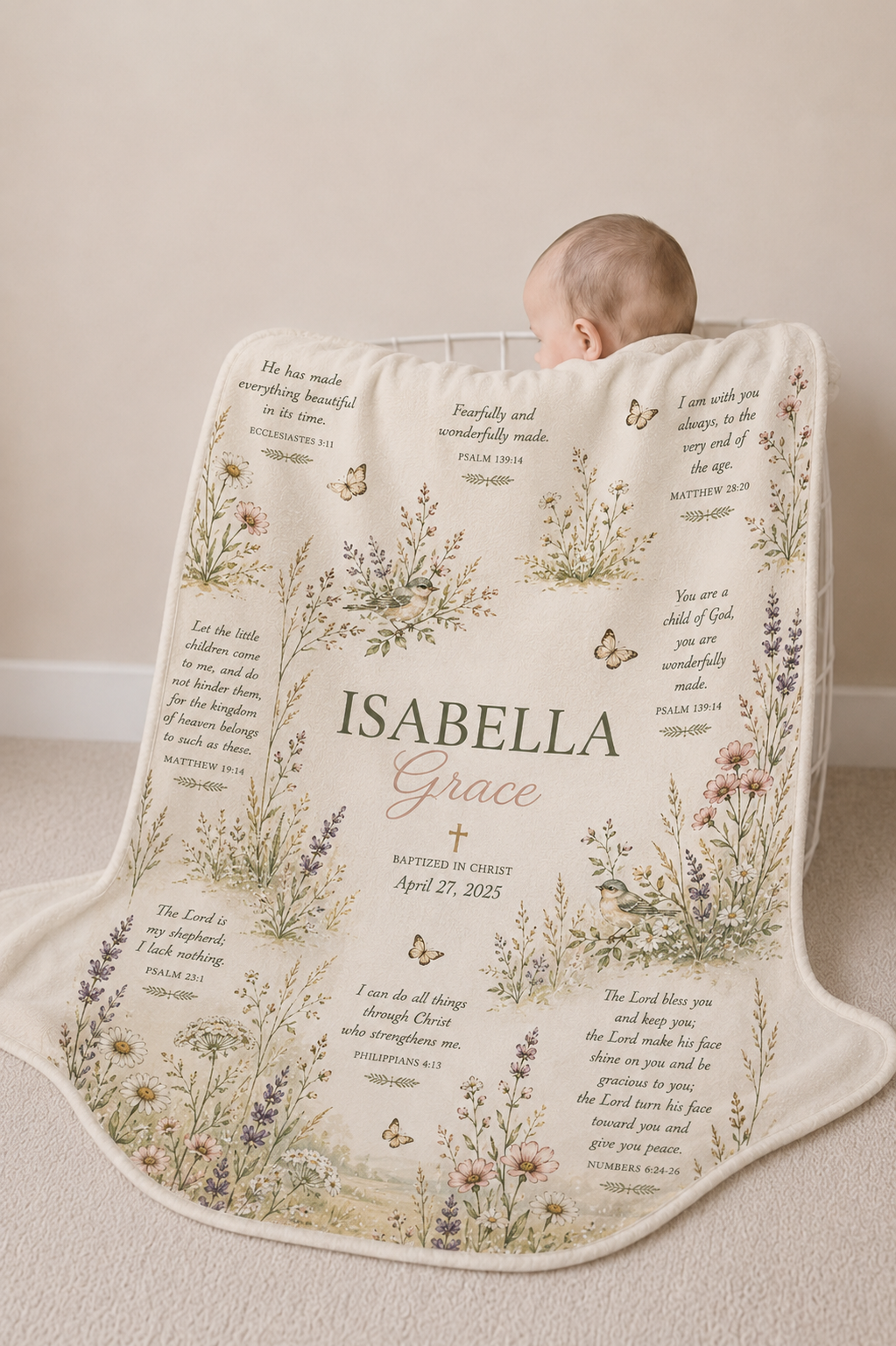 A baby is lying on a personalized cream-colored blanket with botanical illustrations, cursive text reading 'ISABELLA Grace,' and Bible verses printed around the border. The baby is photographed in a soft neutral indoor setting.