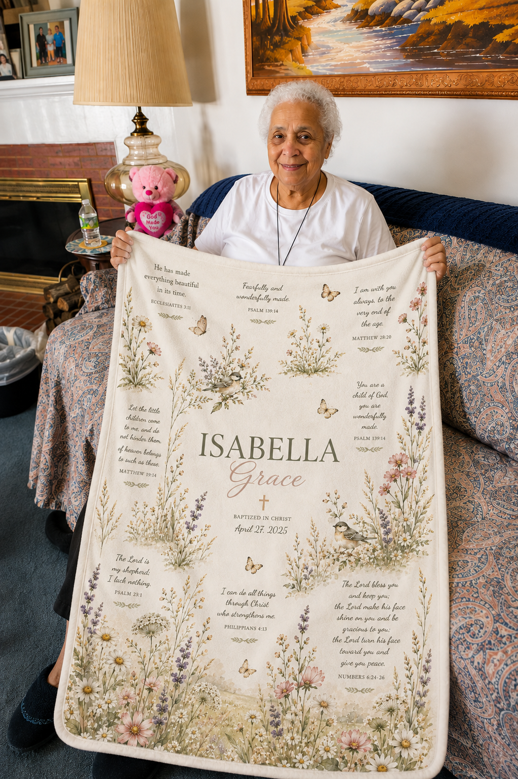 An elderly woman sits on a couch holding up a large personalized memorial blanket. The blanket features a cream background decorated with delicate botanical illustrations, butterflies, and printed words and phrases, with the name 'ISABELLA Grace' prominently displayed in the center.