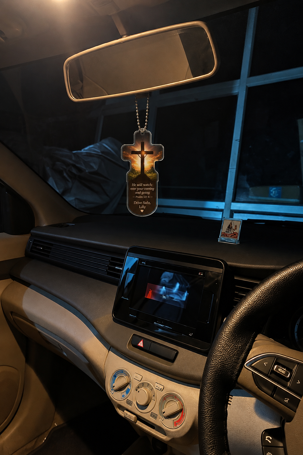 A car interior with a hanging cross-shaped air freshener or ornament featuring a glowing cross design and text, suspended near the rearview mirror above the dashboard.